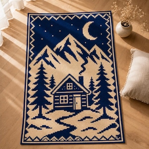 May include: A rectangular woven rug featuring a dark blue and cream pixelated landscape design. The scene depicts a cabin with smoke rising from the chimney, surrounded by pine trees, snow-covered ground, and mountains under a crescent moon.