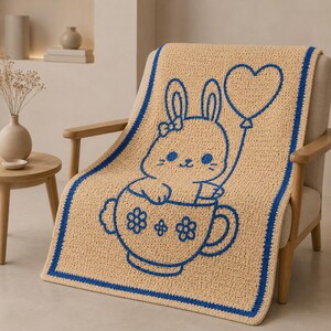 May include: A beige crochet blanket with a blue border and a blue cartoon bunny design. The bunny is in a teacup and holding a heart-shaped balloon. The blanket is draped over a wooden chair.