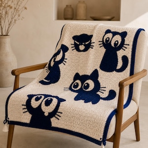 May include: Cream-colored crochet blanket featuring navy blue cat designs. The blanket is draped over a chair with wooden arms. The cats have large eyes and various poses. A navy blue border and tassels complete the design.