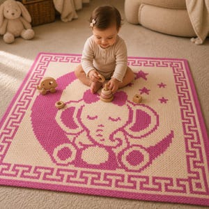 May include: A pink and cream baby blanket featuring an elephant design on a crescent moon. The blanket has a geometric border and is surrounded by wooden toys. The blanket is made of soft material.