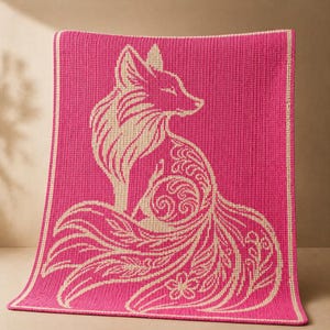 May include: A hot pink knitted blanket featuring a cream-colored fox design. The fox is depicted in a stylized, decorative manner with intricate floral and swirl patterns. The blanket has a cream-colored border.