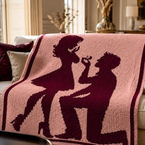 Pode incluir: Um cobertor de malha rosa com uma silhueta bord&ocirc; de um pedido de casamento. O design mostra uma pessoa ajoelhada oferecendo um anel a outra pessoa. A borda e as silhuetas s&atilde;o de cor bord&ocirc; escuro.