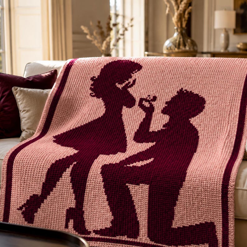Pode incluir: Um cobertor de malha rosa com uma silhueta bord&ocirc; de um pedido de casamento. O design mostra uma pessoa ajoelhada oferecendo um anel a outra pessoa. A borda e as silhuetas s&atilde;o de cor bord&ocirc; escuro.