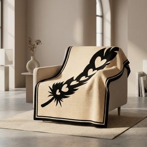 May include: A cream-coloured throw blanket with a black feather design featuring heart shapes. The blanket has a black border and is draped over a beige armchair. The design is a striking contrast against the neutral background.