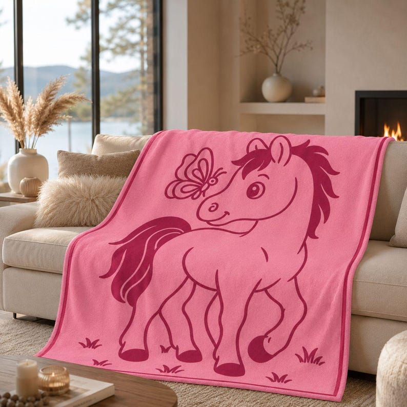 May include: A soft pink fleece blanket featuring a dark pink graphic illustration of a pony standing in a field with a butterfly near its head. The blanket has a solid pink border and small dark pink grass motifs along the bottom edge.