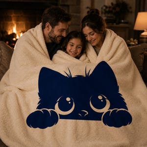May include: A cream-colored sherpa blanket featuring a navy blue cat face design. The blanket is wrapped around a family of three, creating a cozy and warm scene. The cat design has large eyes and paws.