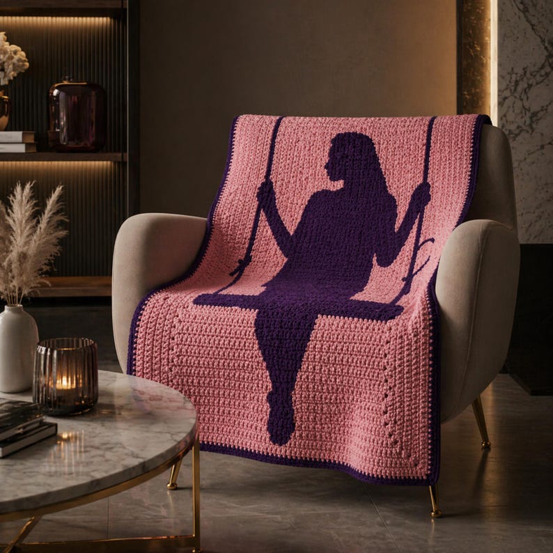 Puede incluir: Una manta rosa tejida a ganchillo con una silueta p&uacute;rpura oscura de una persona en un columpio. La manta est&aacute; sobre un sill&oacute;n beige. La manta tiene un borde p&uacute;rpura oscuro.