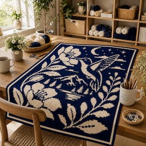 May include: A rectangular table runner featuring a dark blue background with a cream-colored woven design of a hummingbird, large flowers, mountains, and a crescent moon. The textile has a distinct grid-like texture and a solid border frame.