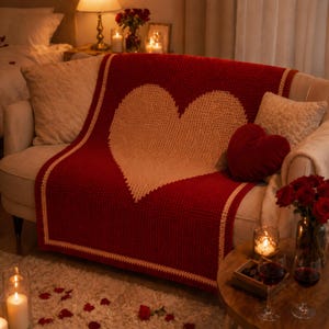 May include: A red knitted throw blanket with a large cream heart design on a beige sofa. The blanket has a cream border. A red heart-shaped cushion and candles create a cosy, romantic atmosphere. The scene is lit by warm, soft light.