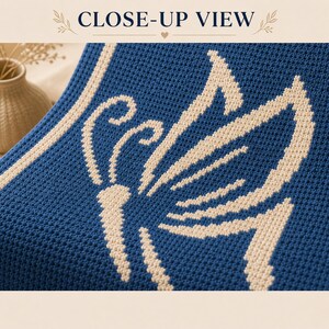 May include: Close-up of a blue crocheted blanket featuring a cream-coloured butterfly design. The blanket has a textured appearance and a cream-coloured border. The words "CLOSE-UP VIEW" are at the top of the image.