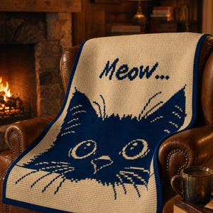 May include: A beige and navy blue crocheted throw blanket featuring a cat face design and the word "Meow...". The blanket is draped over a brown leather armchair. A mug of coffee sits on a wooden table next to the chair.