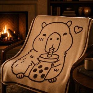 May include: A tan throw blanket draped over a wooden chair. The blanket features a brown cartoon illustration of a capybara drinking from a boba tea cup, with a heart symbol. The background includes a fireplace and a side table.