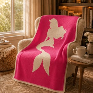 May include: A hot pink throw blanket with a cream-colored mermaid silhouette design. The blanket is draped over a beige armchair. The blanket has a cream-colored border and is made of a soft material.