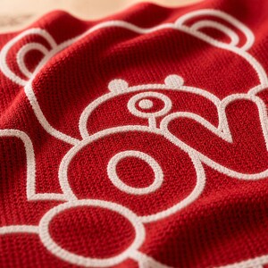 May include: Close-up of a red towel with a white outline of a bear design. The towel has a textured surface and is folded, showing the bear's head and the number 2. The design is a playful and modern style.