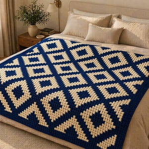 May include: A navy blue and cream-coloured crochet blanket with a geometric diamond pattern. The blanket is draped over a bed with neutral-toned pillows and a beige bedspread. The blanket's design features repeating diamond shapes.