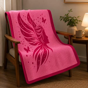 May include: A pink knitted blanket with a fairy design draped over a wooden chair. The blanket features a dark pink border and a central design of a fairy silhouette with wings and butterflies. The blanket is made of soft material.