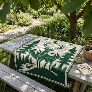 Peut inclure: Une couverture carr&eacute;e au crochet pr&eacute;sentant un motif vert fonc&eacute; et cr&egrave;me repr&eacute;sentant un cerf dans une for&ecirc;t avec des pins, des montagnes, un croissant de lune et des &eacute;toiles. La couverture a une bordure simple sur une table en bois.