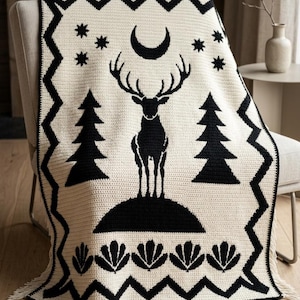 K&ouml;nnte beinhalten: Eine cremefarbene H&auml;keldecke mit einem schwarzen grafischen Design, das einen stehenden Hirsch, Tannenb&auml;ume, eine Mondsichel und Sterne zeigt. Die Decke hat einen schwarzen Zickzack-Rand und eine Reihe schwarzer f&auml;cherf&ouml;rmiger Motive am unteren Ende.