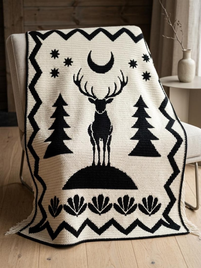 K&ouml;nnte beinhalten: Eine cremefarbene H&auml;keldecke mit einem schwarzen grafischen Design, das einen stehenden Hirsch, Tannenb&auml;ume, eine Mondsichel und Sterne zeigt. Die Decke hat einen schwarzen Zickzack-Rand und eine Reihe schwarzer f&auml;cherf&ouml;rmiger Motive am unteren Ende.