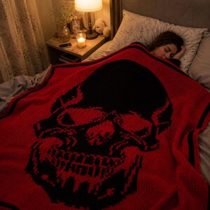 Puede incluir: Una manta roja y negra con un patr&oacute;n de calavera cubre a una persona durmiendo en la cama. La manta presenta un gran dise&ntilde;o de calavera negra sobre un fondo rojo. La habitaci&oacute;n est&aacute; tenuemente iluminada, con una mesita de noche y una l&aacute;mpara.