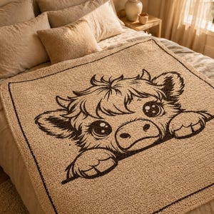 May include: A beige throw blanket featuring a cartoon cow face in dark brown. The cow has large eyes and is peeking over the edge. The blanket is on a bed with beige pillows. The blanket's dimensions are approximately 150 cm x 150 cm.