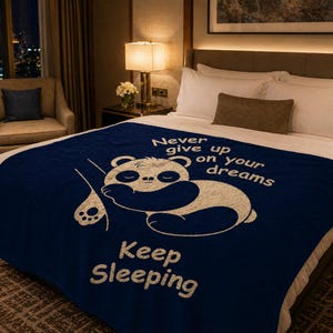 May include: A navy blue blanket featuring a white panda design and the text "Never give up on your dreams" and "Keep Sleeping". The blanket is on a bed with white pillows and a brown cushion.