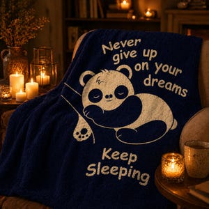 May include: A navy blue throw blanket featuring a white panda bear design and the text "Never give up on your dreams, Keep Sleeping." The blanket is on a chair, with candles creating a warm, inviting atmosphere in the background.