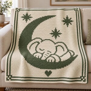 May include: A cream-colored crochet blanket featuring a dark green design of a sleeping elephant resting on a crescent moon. The pattern includes three dark green stars and a small heart at the bottom, all framed by a dark green geometric border.