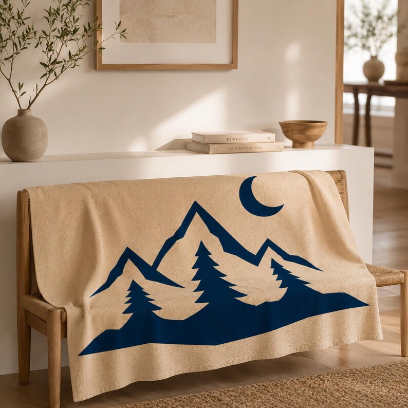 May include: A beige throw blanket featuring a dark blue graphic print of a mountain range, evergreen trees, and a crescent moon. The blanket is draped over a wooden bench, displaying a clean, high-contrast design with sharp geometric shapes.