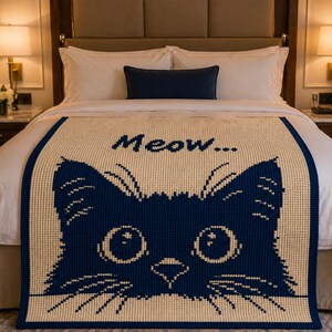 May include: A cream-colored throw blanket with a navy blue cat face design and the word "Meow..." in navy blue. The blanket is placed on a bed with white linens and a navy blue pillow.