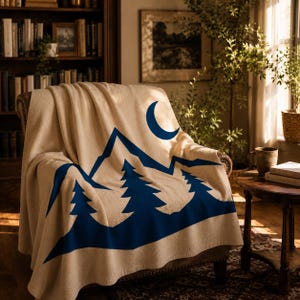 May include: A beige throw blanket draped over a chair, featuring a bold navy blue graphic print of a mountain range, three evergreen trees, and a crescent moon. The fabric has a soft, textured appearance with clean, sharp lines in the design.