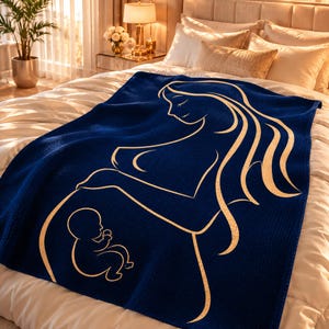 May include: A navy blue blanket with a gold outline depicting a pregnant person and a baby. The blanket is on a bed with white bedding. The design is a simple, elegant illustration.