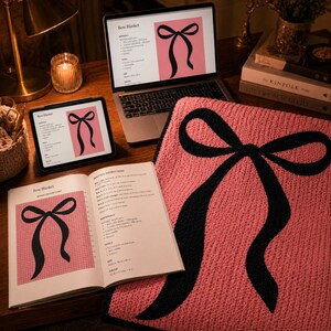 Pu&ograve; includere: Una coperta rosa lavorata a maglia con un grande disegno a fiocco nero, esposta con un laptop, un tablet e un libro aperto che mostra il motivo. Gli schermi del laptop e del tablet mostrano le parole "Bow Blanket". Sono visibili anche una candela accesa e una lampada.
