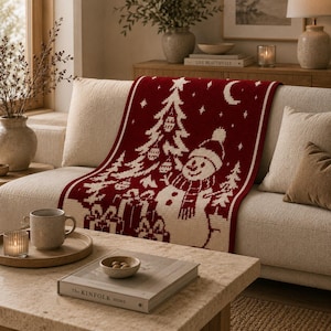 May include: A rectangular knit throw blanket featuring a deep red background with a white pattern depicting a snowman wearing a hat and scarf, a decorated Christmas tree, wrapped gift boxes, a crescent moon, and scattered stars.