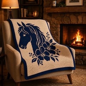 May include: A beige throw blanket with a navy blue horse head and floral design. The blanket has a navy blue border and is draped over a beige armchair. The background includes a fireplace and a lamp.