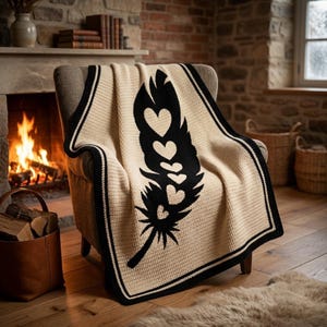 May include: A cream-coloured throw blanket with a black feather design featuring heart cutouts is draped over a chair. The blanket has a black border and is placed in front of a fireplace. The room has a cosy, warm atmosphere.