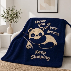 May include: A navy blue throw blanket featuring a sleeping panda bear illustration and the text "Never give up on your dreams, Keep Sleeping" in cream. The blanket is draped over a white armchair.