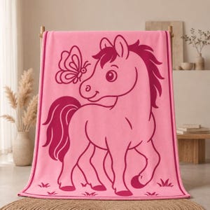 May include: Pink beach towel with a cartoon pony and butterfly design. The pony is outlined in a darker pink, with a matching mane and tail. The butterfly is also outlined in the darker pink. The towel is hanging on a wooden rack.
