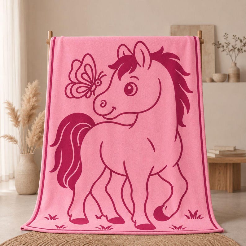 May include: Pink beach towel with a cartoon pony and butterfly design. The pony is outlined in a darker pink, with a matching mane and tail. The butterfly is also outlined in the darker pink. The towel is hanging on a wooden rack.