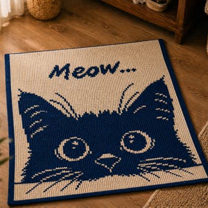 May include: A rectangular, cream-colored rug with a navy blue cat face design. The cat's eyes and nose are detailed with white accents. The word "Meow..." is written above the cat's head in navy blue. The rug has a navy blue border.