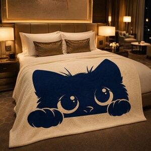 May include: A cream-colored blanket with a large navy blue cat face design. The cat's eyes are large and round, and its paws are visible at the bottom. The blanket is on a bed with white pillows and a brown headboard.