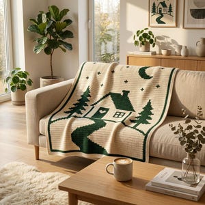 Pode incluir: Uma manta de croch&ecirc; na cor creme com um design gr&aacute;fico em verde escuro de uma pequena casa com um cora&ccedil;&atilde;o na porta, rodeada por pinheiros, um caminho sinuoso, uma lua crescente e pequenas estrelas, emoldurada por uma borda verde s&oacute;lida.