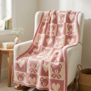 May include: A pink and cream crocheted blanket with a pattern of teddy bears and hearts, draped over a white rocking chair. The blanket is made of soft yarn and is suitable for a nursery or as a decorative throw. The blanket is in a square pattern.