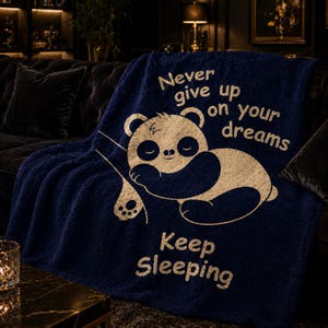 May include: A navy blue throw blanket featuring a sleeping panda bear design and the text "Never give up on your dreams" and "Keep Sleeping" in white. The blanket is made of a soft, plush material.