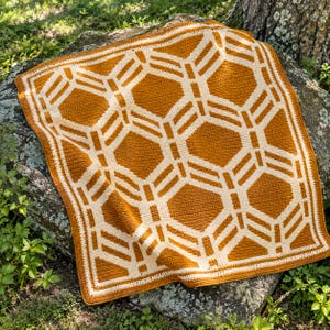 May include: A square crochet blanket featuring a geometric pattern of interlocking hexagons and lines in mustard yellow and cream white. The textile displays a consistent stitch texture throughout, with a solid border framing the design.