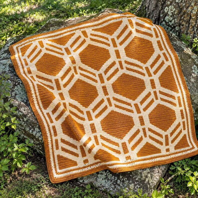 May include: A square crochet blanket featuring a geometric pattern of interlocking hexagons and lines in mustard yellow and cream white. The textile displays a consistent stitch texture throughout, with a solid border framing the design.