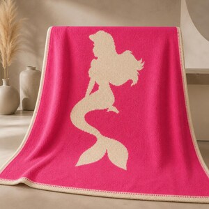 May include: A hot pink blanket with a cream-colored mermaid silhouette. The blanket has a cream-colored border. The mermaid design features a long flowing tail and hair. The blanket is folded over a surface.