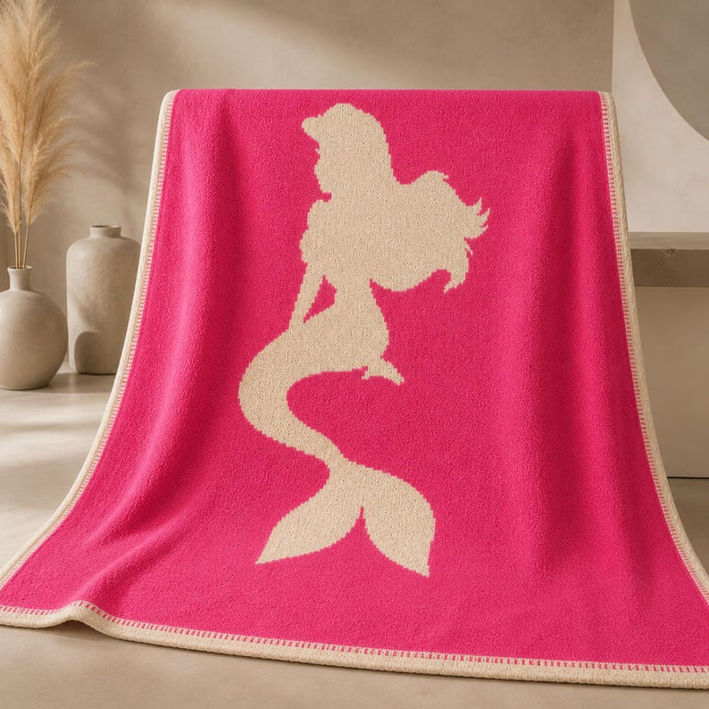 May include: A hot pink blanket with a cream-colored mermaid silhouette. The blanket has a cream-colored border. The mermaid design features a long flowing tail and hair. The blanket is folded over a surface.