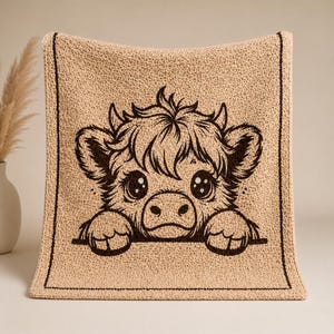 May include: A beige knitted blanket with a cartoon Highland cow design. The cow's face is depicted in dark brown, with large eyes and a friendly expression. The blanket has a dark brown border and a textured appearance.