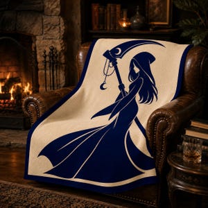 May include: A rectangular throw blanket with a cream-coloured base and a dark navy blue graphic print. The design features a hooded figure in a long flowing gown holding a large scythe with a crescent moon charm hanging from the handle.